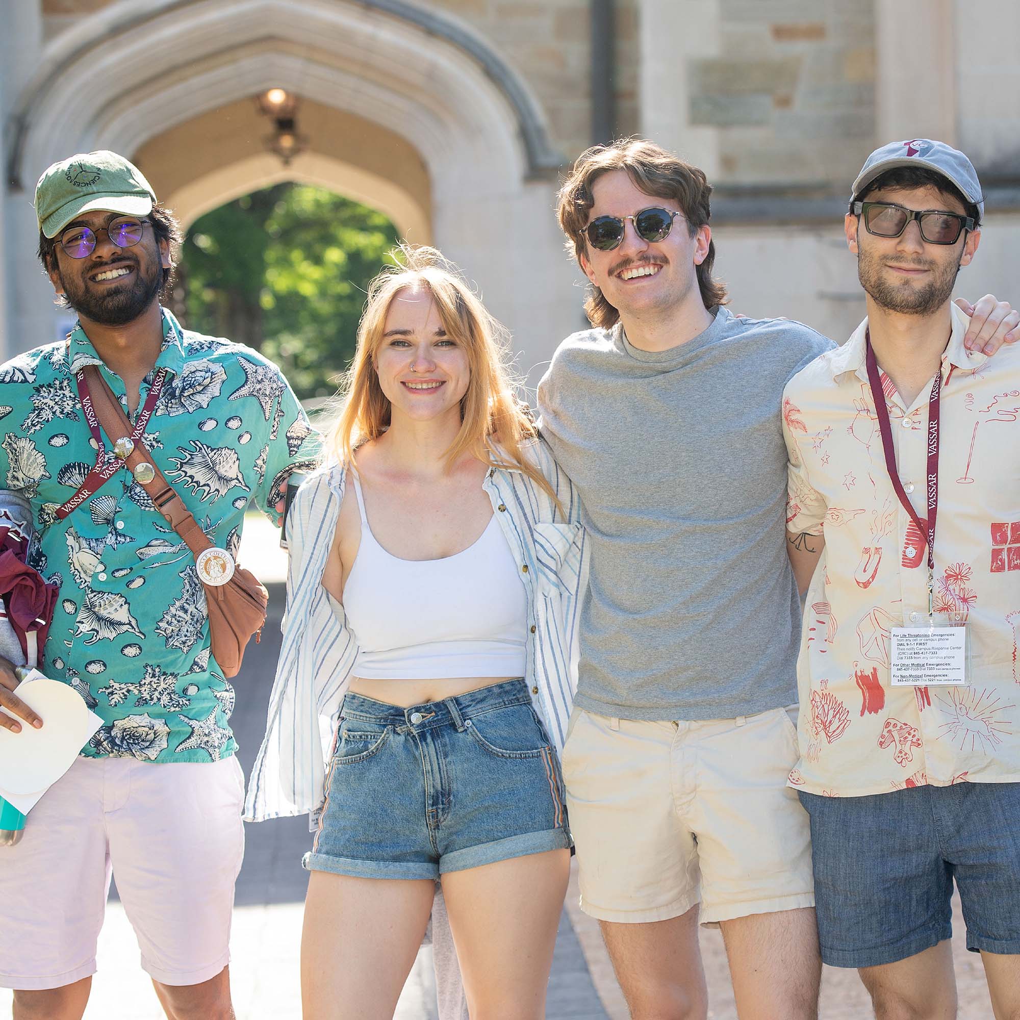 Four people stand with their arms around one another in summer clothing on a sunny day in front of Vassar’s Main Gate.